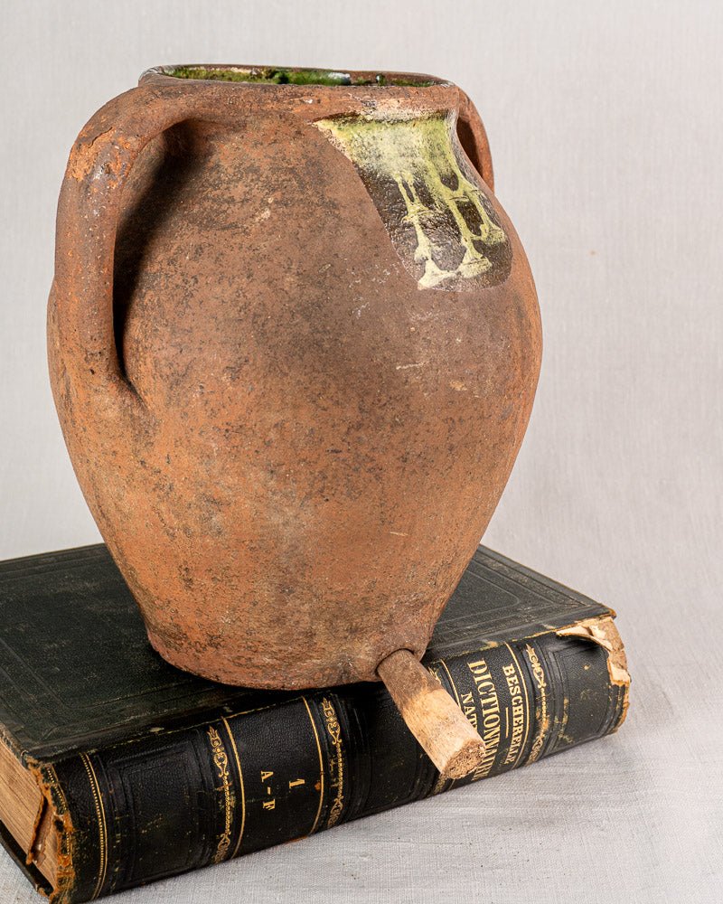 Antique French glazed terracotta vinaigrier with chipped spout and mossy stains on vintage books.