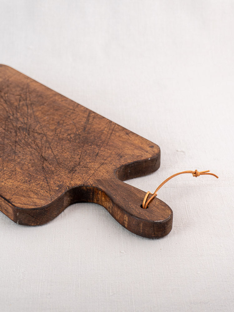 Antique French cutting board with paddle handle, rectangular form, visible scratches, and leather loop on light fabric background.