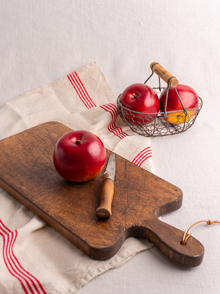 Antique French cutting board with paddle handle, red apple, knife, and wire basket of apples on striped cloth.