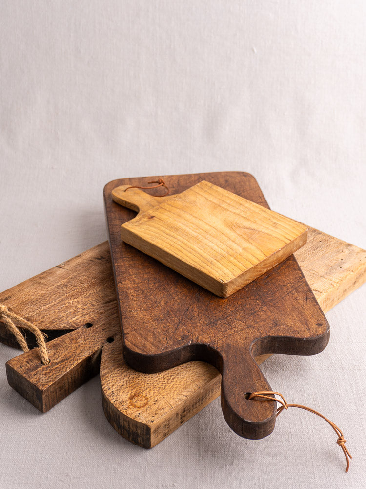Antique French cutting boards with paddle handles and leather loops, stacked on a light fabric surface.
