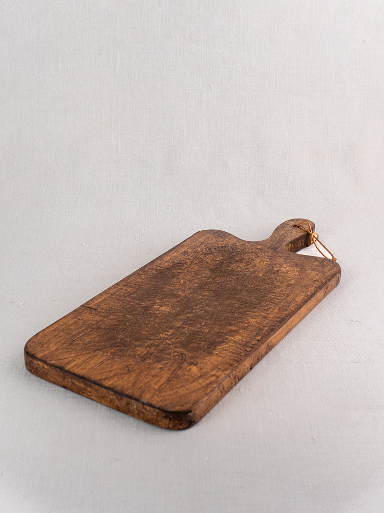 Antique French cutting board with paddle handle and metal loop, rectangular shape, on a textured light surface.