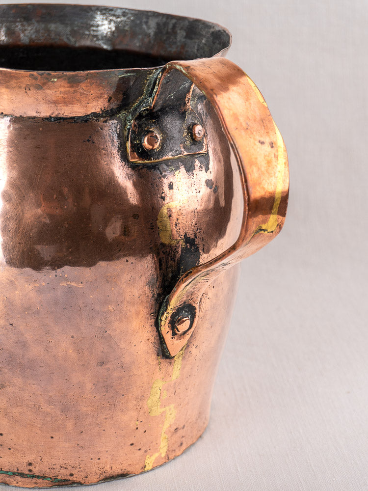 Antique French hammered copper pitcher with large riveted handle, soldered seams, visible repairs, and patinated surface.