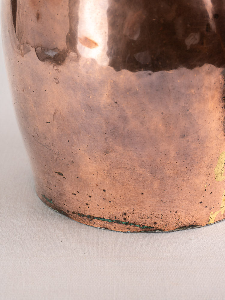 Lower section of antique French hammered copper pitcher with greenish patina near base, on light textured surface.