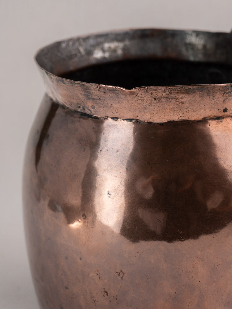 Antique French copper pitcher with a wide, uneven hammered rim and aged patina, shown on a softly blurred background.