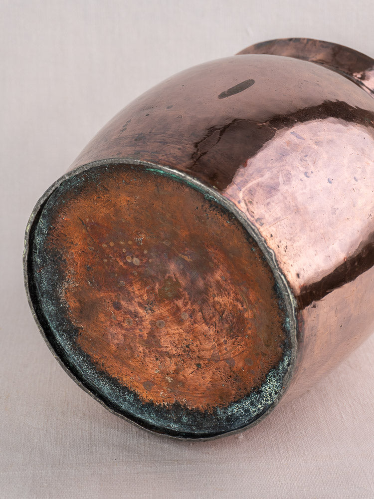 Antique French hammered copper pitcher with oxidized, weathered base, shown resting on a neutral surface.
