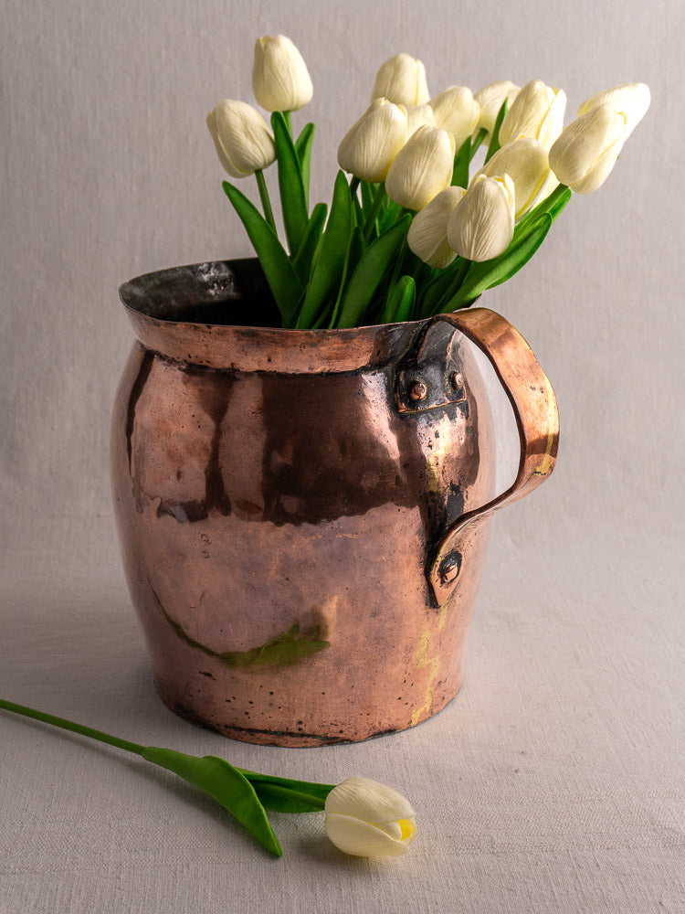 Antique French hammered copper pitcher with handle, filled with white tulips, on light fabric and neutral background.