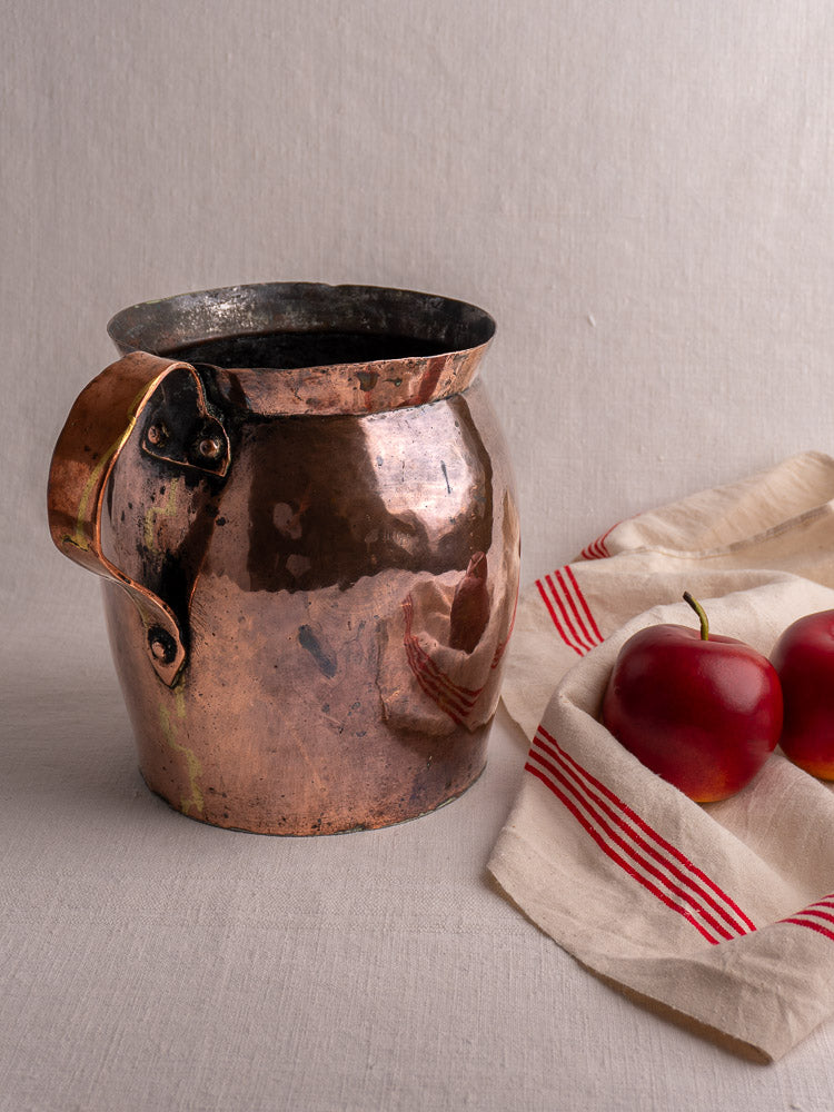 Antique French hammered copper pitcher with handle on beige fabric, beside white cloth with red stripes and two red apples.