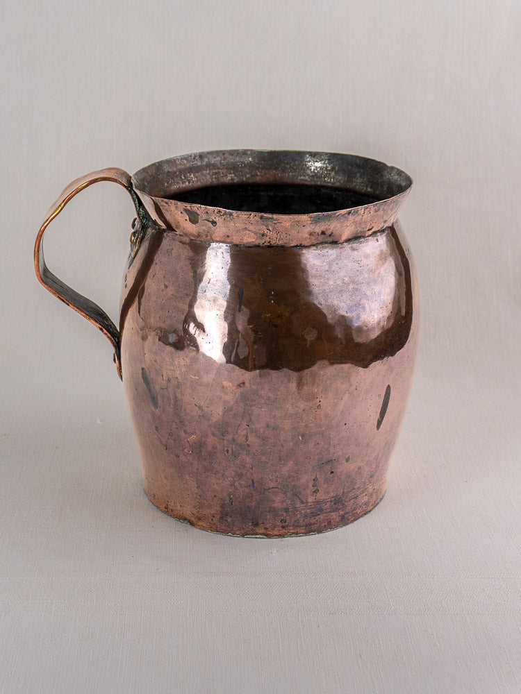 Antique French hammered copper pitcher with wide mouth, curved handle, and patina, set on a light gray background.