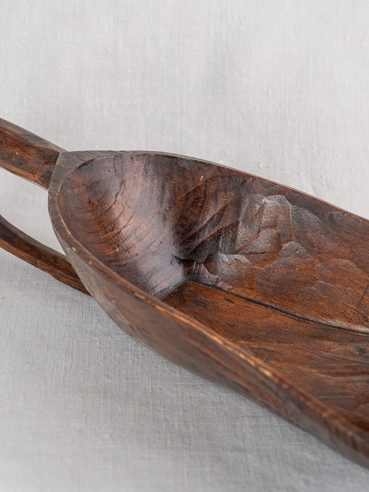 Antique French wooden grain scoop with rich dark wood grain and curved interior, shown on a light fabric background.