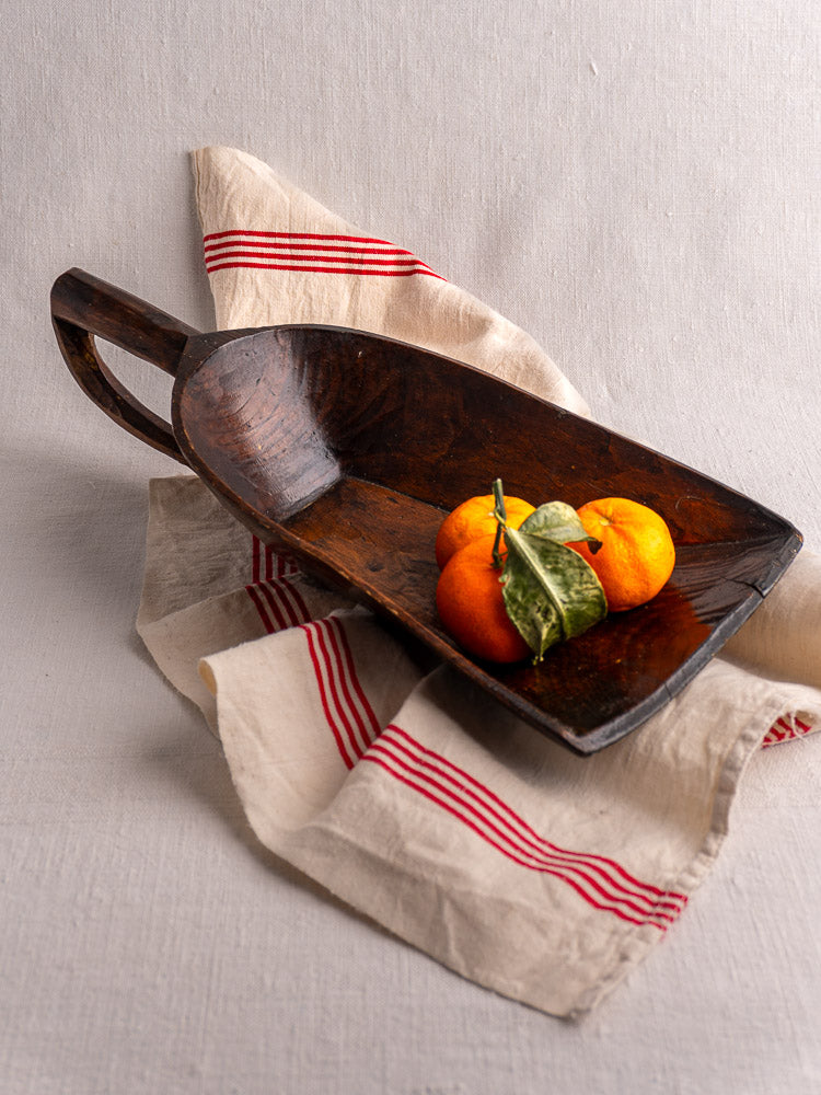 Antique French wooden grain scoop with three oranges on beige cloth with red stripes, neutral backdrop.