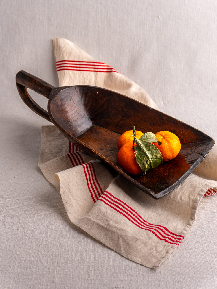 Antique French wooden grain scoop holding three citrus fruits with leaves on beige cloth with red stripes.