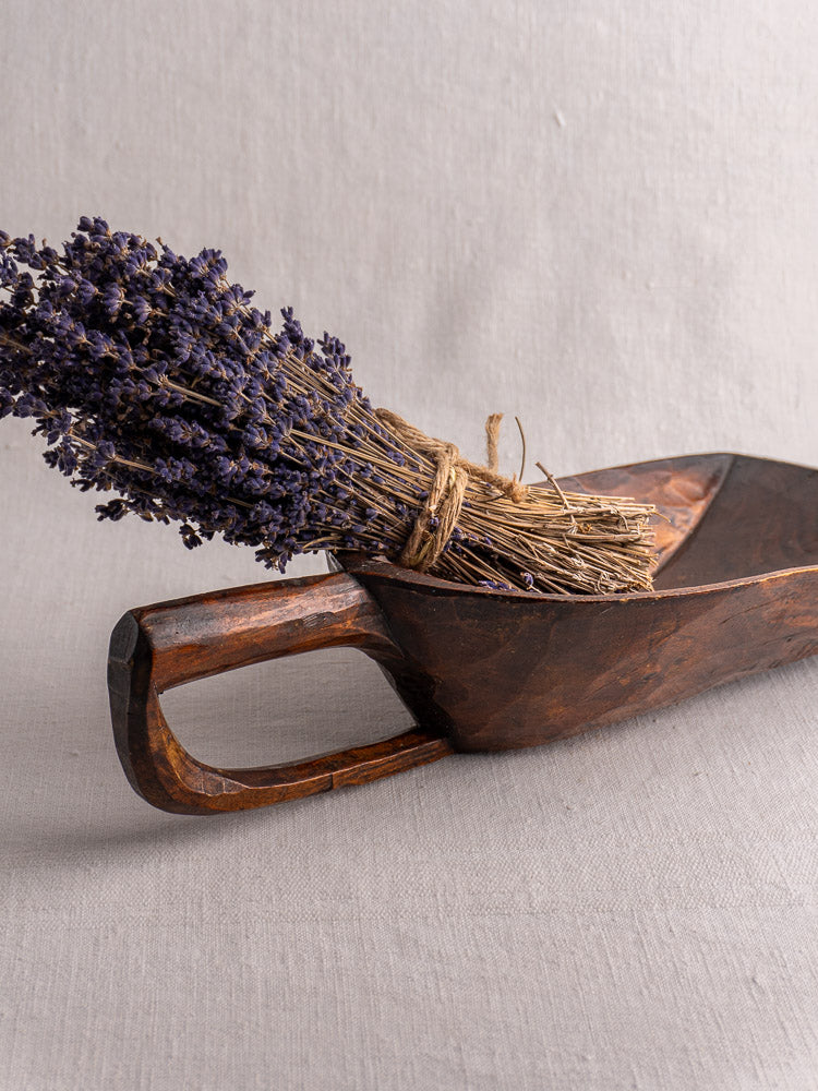 Antique French hand carved 19th-century wooden grain scoop holding dried lavender, on a neutral fabric background.
