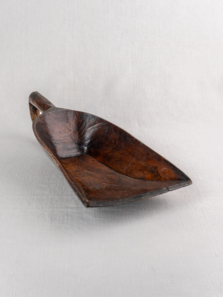 Antique French wooden grain scoop with rectangular curved bowl and short handle on light grey fabric background.