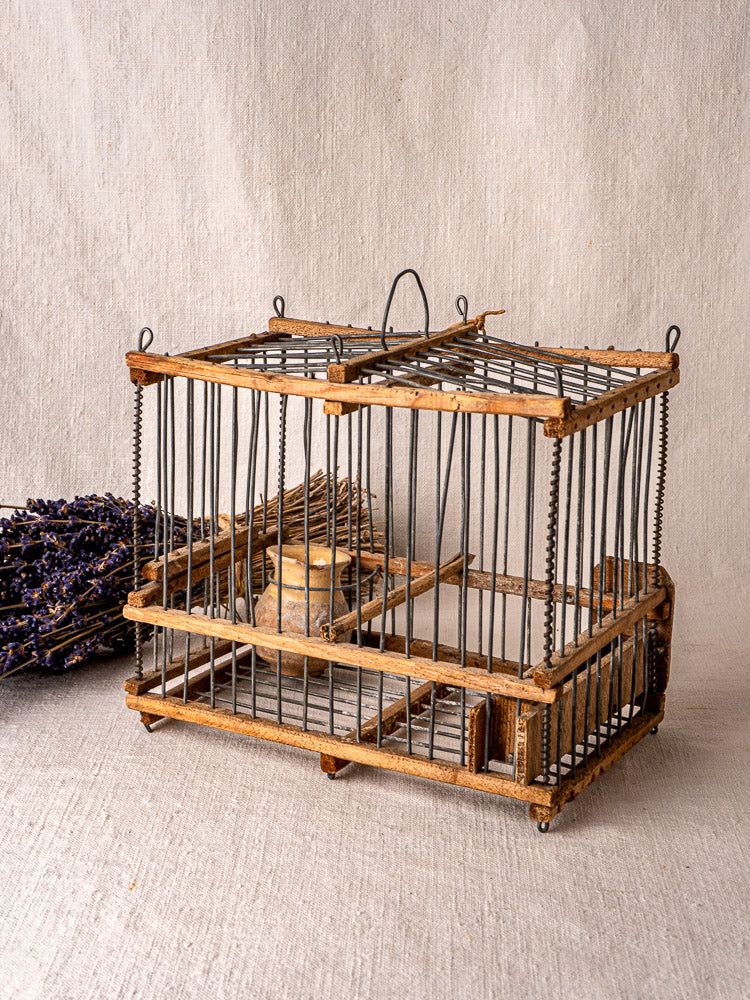 Antique French folk art bird cage in wood and wire with ceramic dish, displayed on beige fabric, dried lavender behind.