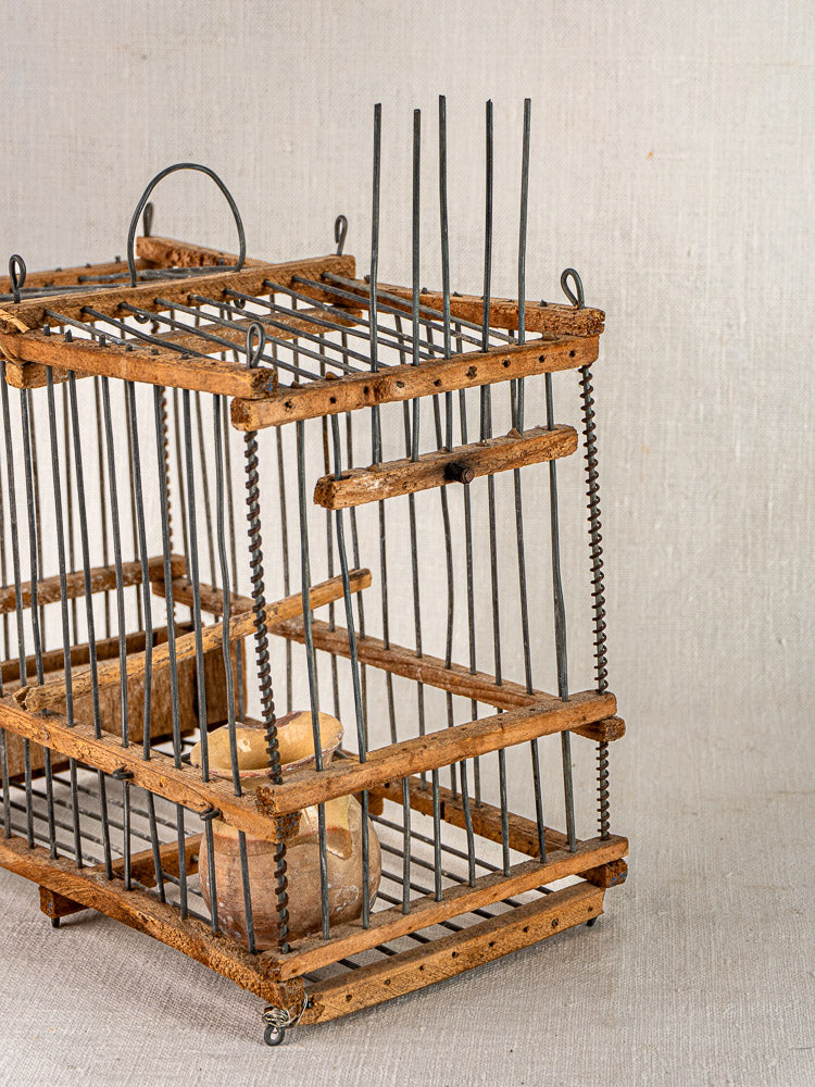 Antique French folk art wood and wire bird cage with small clay vessel, set on neutral fabric background.