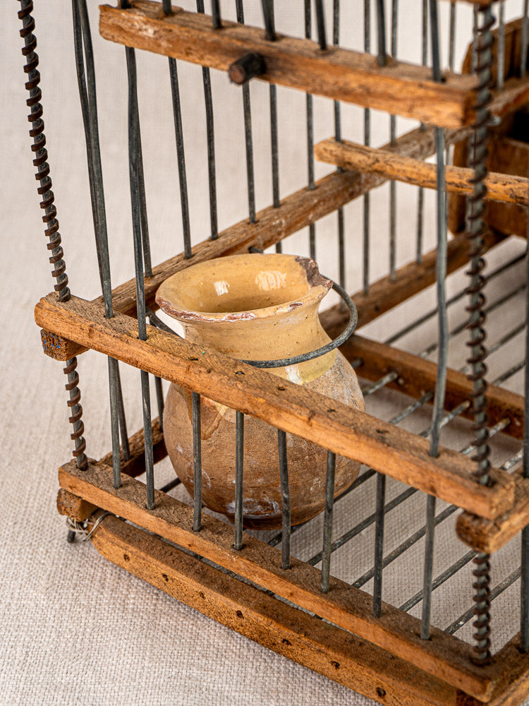 Antique French wooden and wire bird cage with small glazed ceramic jug, displayed on beige fabric.