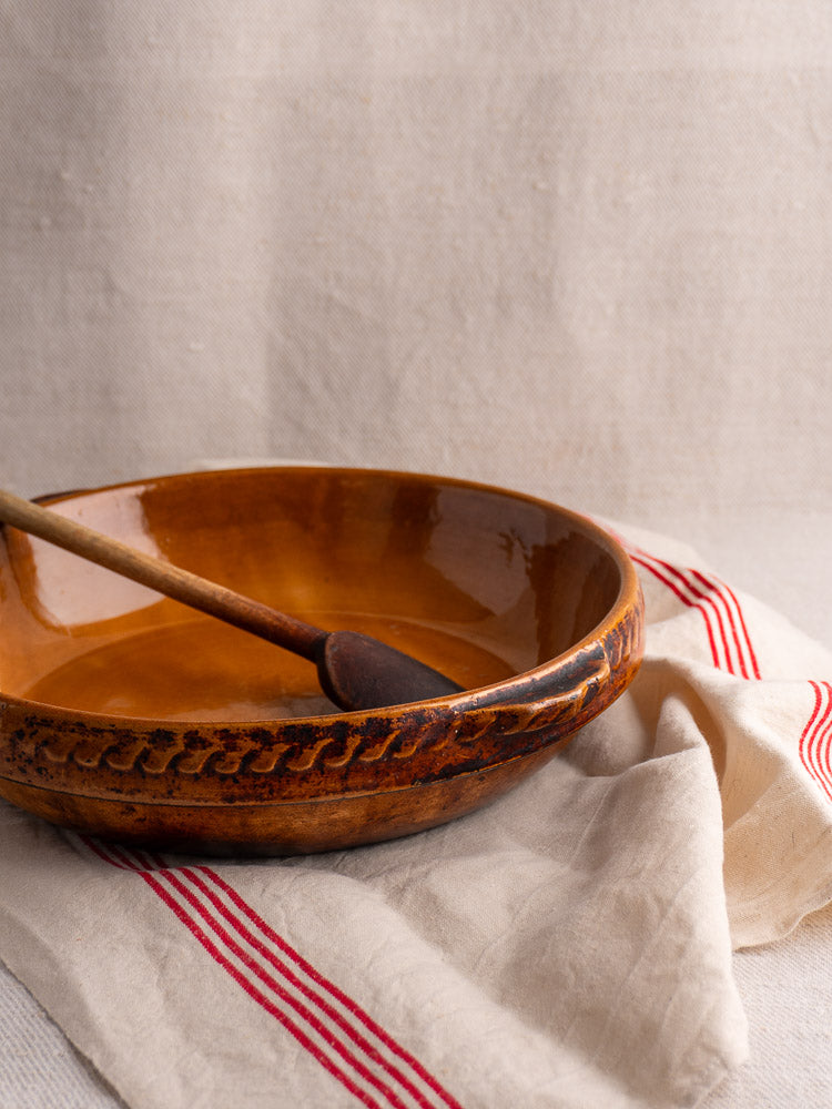 Antique French Vitry-le-François brown glazed stoneware gratin dish with wooden spoon on striped cloth.