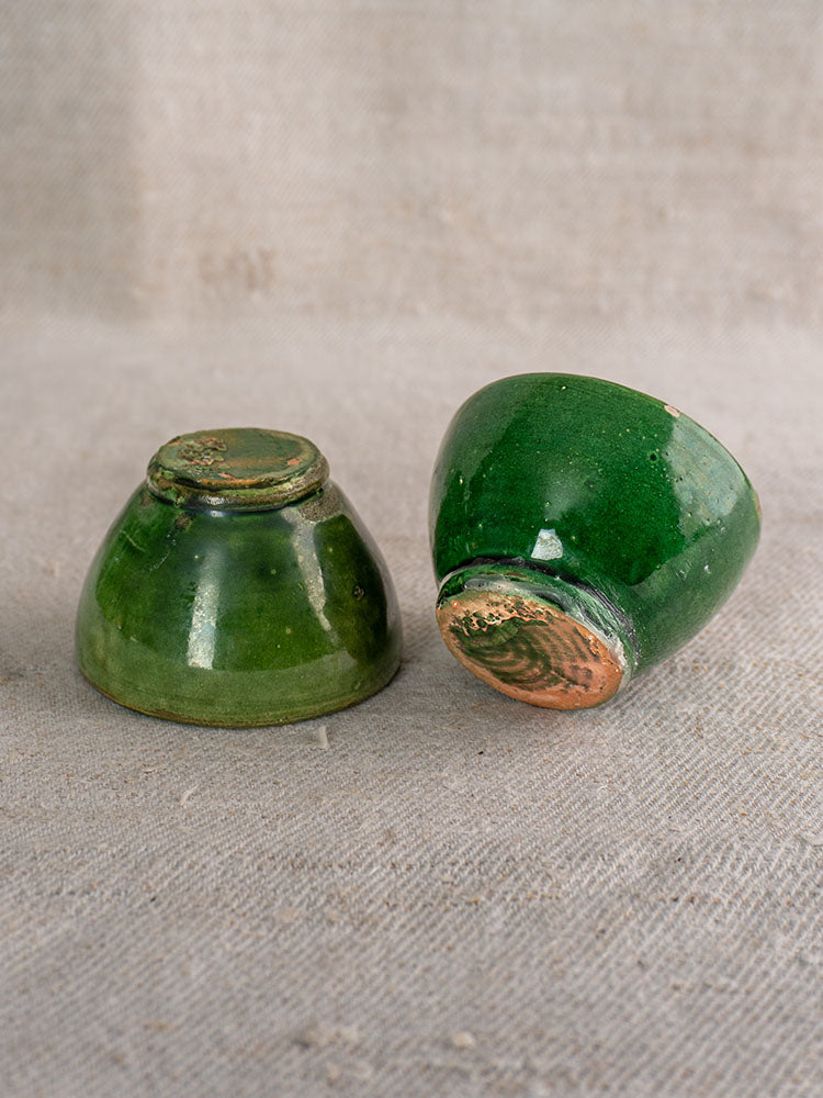 French green glazed ceramic bowls, one upright and one tilted to show base, on textured beige fabric.