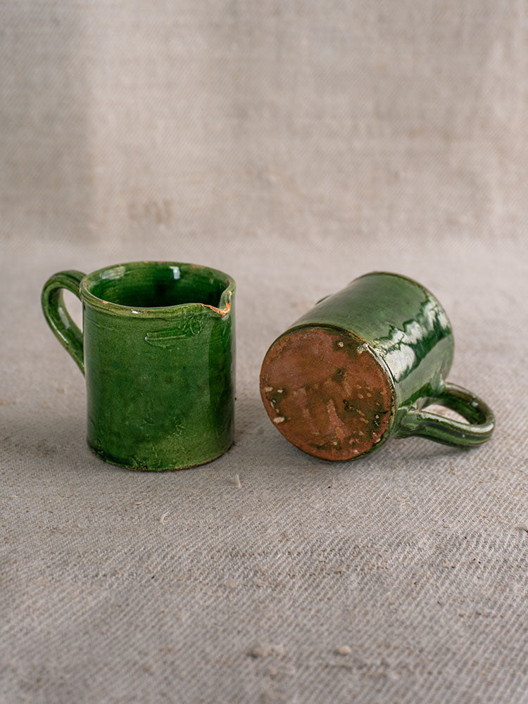 Pair of small French green glazed pitchers, one upright, one on its side showing unglazed base, on neutral textured surface.
