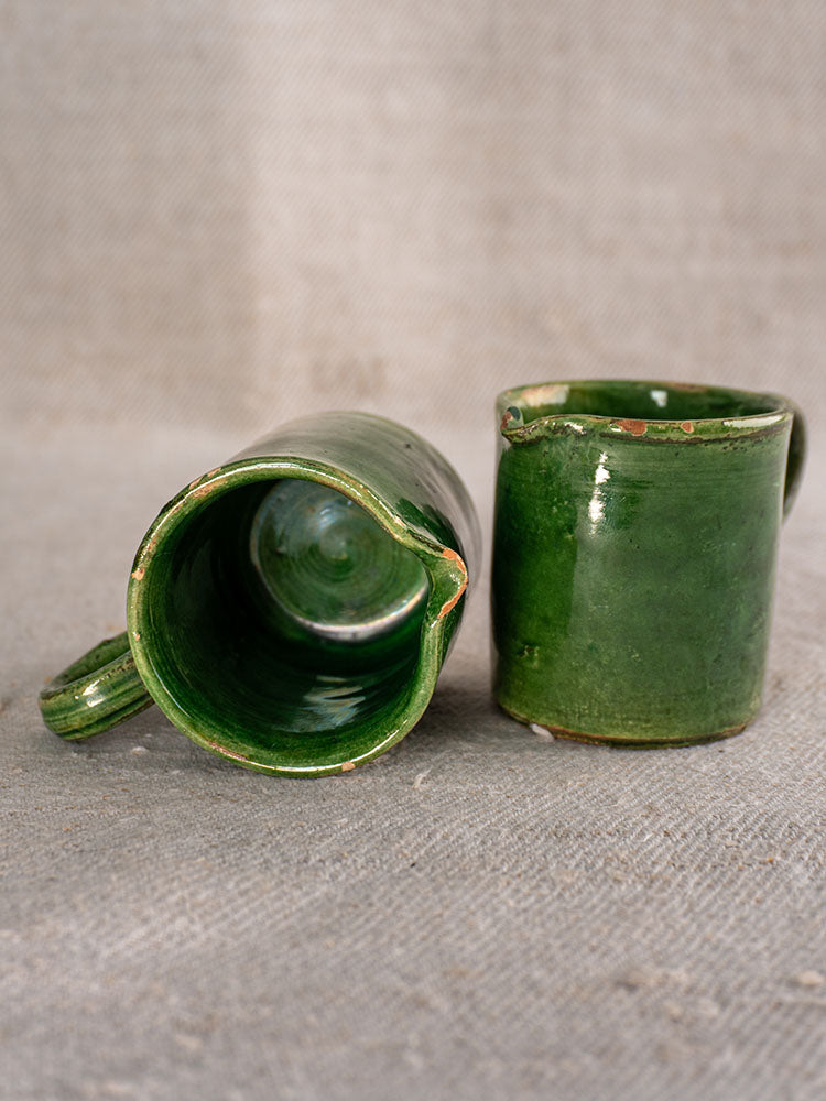 Two French green-glazed ceramic pitchers, one upright, one on its side, displayed on textured beige fabric.