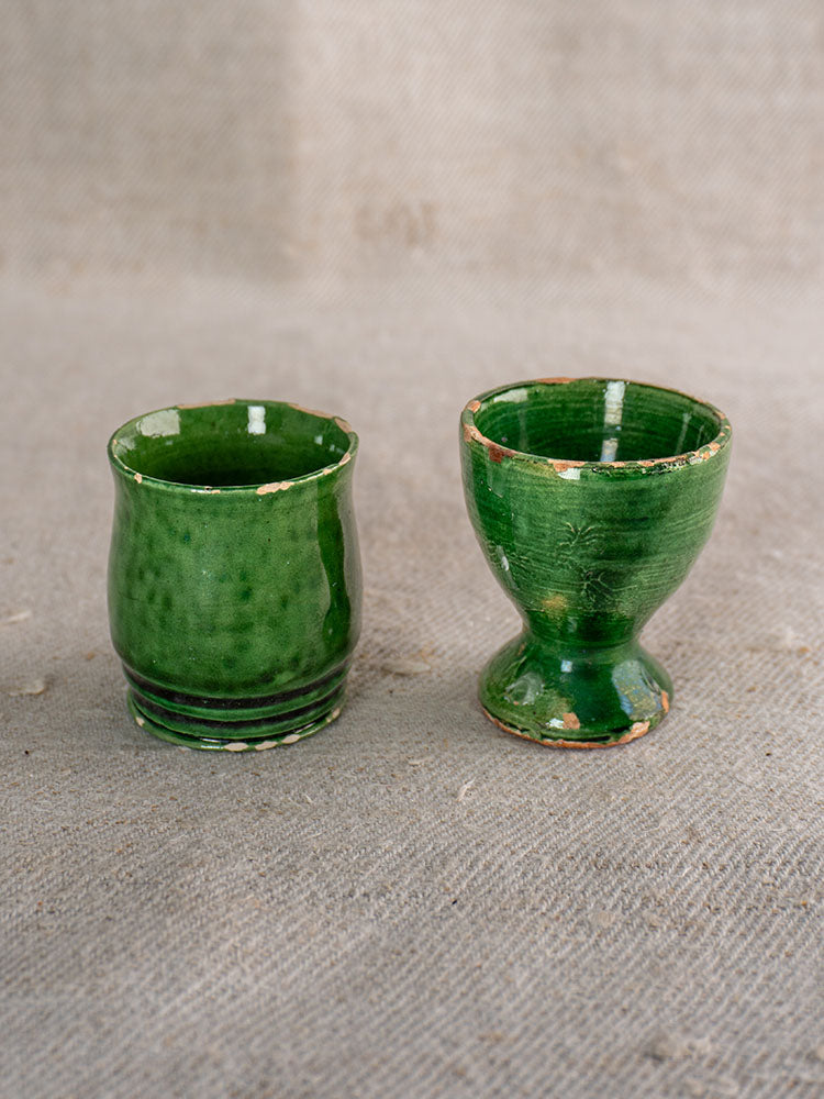 Pair of small green French glazed ceramic cups, straight and flared shapes, on textured gray fabric.