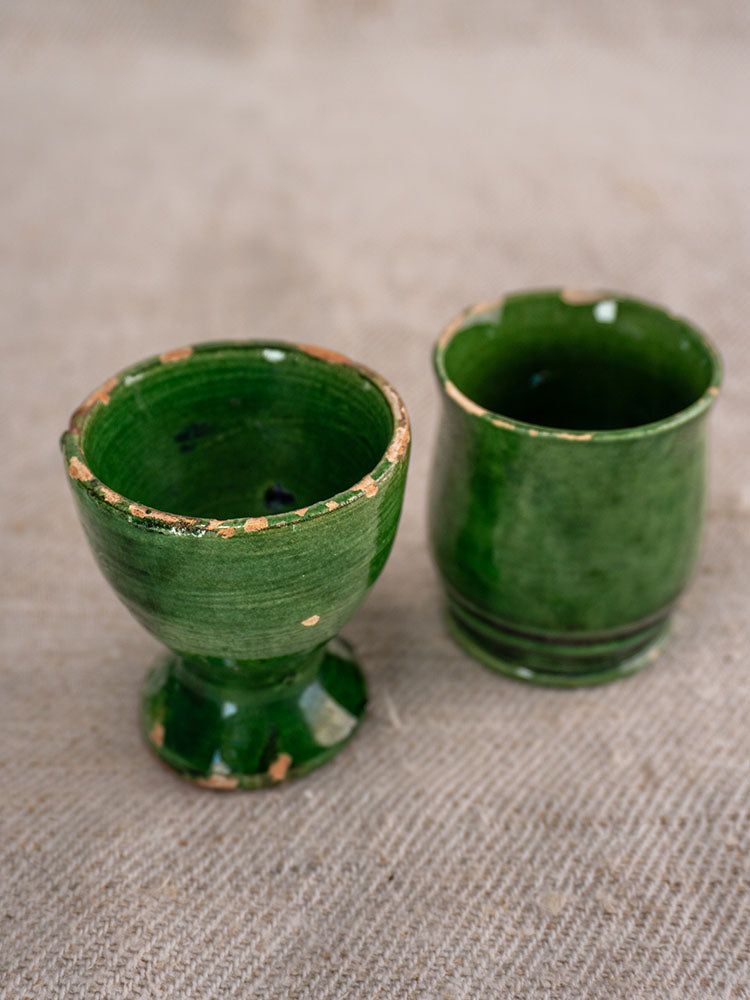 Two small French green glazed cups, one stemmed and one cylindrical with chipped edges, on beige fabric.
