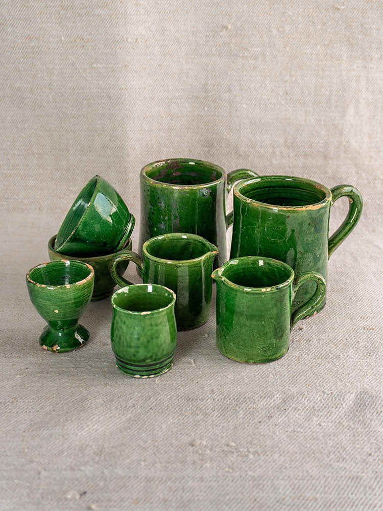 French green glazed Terre Vernissée pottery set of 8 assorted cups and mugs on textured beige fabric background.