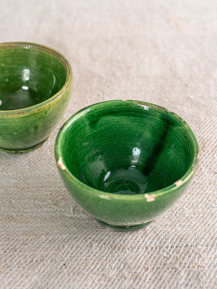 Two small French green glazed pottery bowls with aged, chipped edges on light beige textured fabric.