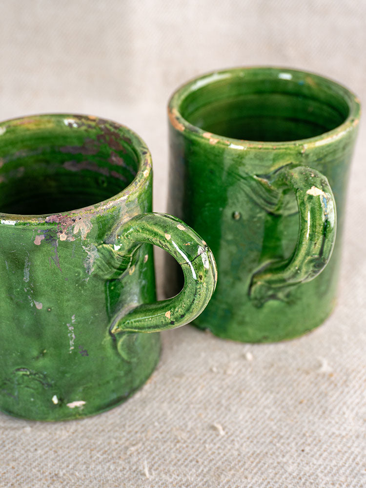 Two vintage French green glazed pottery mugs with large handles and aged glaze on textured beige fabric.