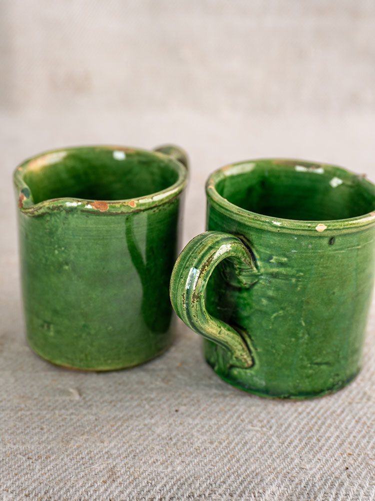 French green glazed pottery cups, one handled and one with spout, show rim chips on beige fabric.