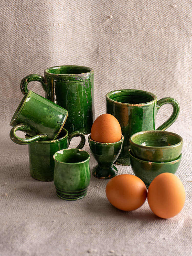 French green glazed Terre Vernissée pottery set of 8 assorted cups and mugs on textured beige fabric background.
