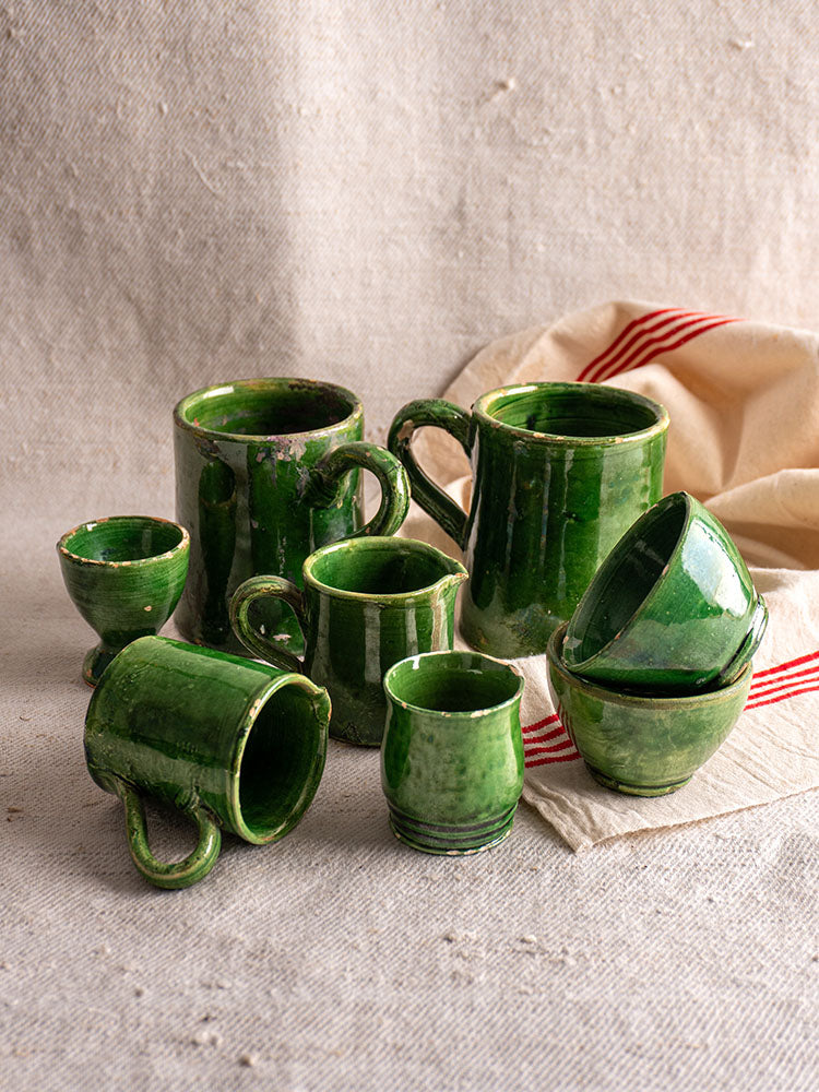 French green glazed pottery 8-piece set displayed on textured beige cloth with striped beige and red napkin in background.