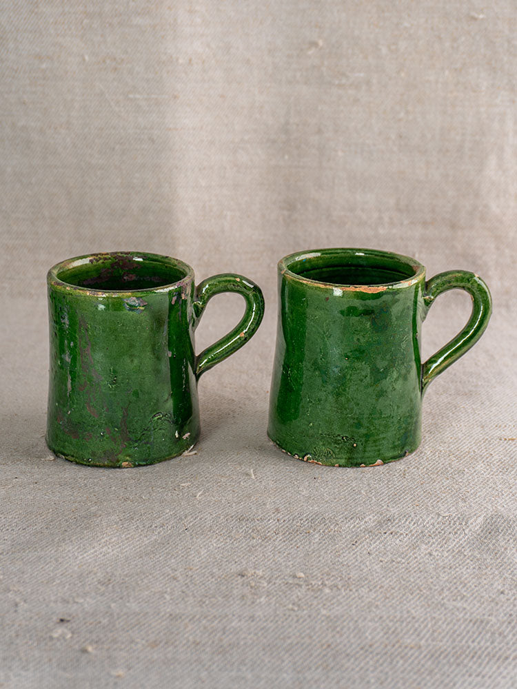 Pair of French green glazed pottery mugs with handmade details from Provençal Terre Vernissée set on beige fabric.