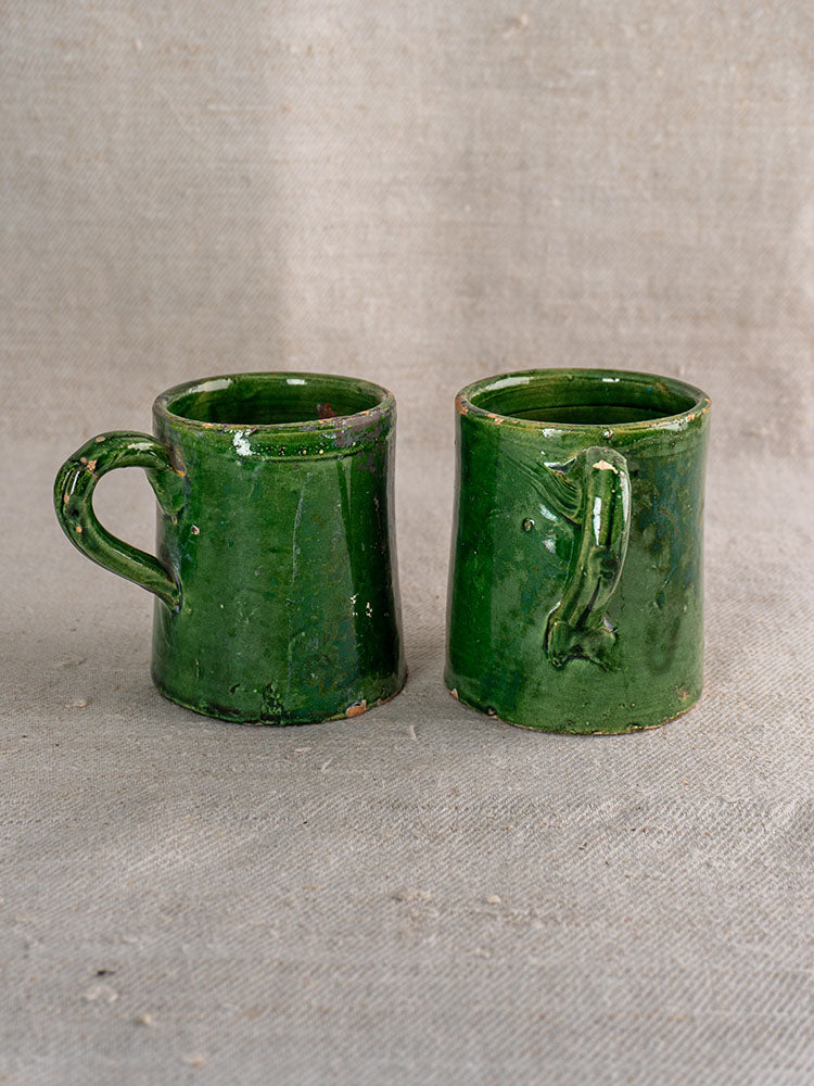 French Green Glazed Pottery mugs with unique handles and rustic glaze, displayed on textured beige fabric.