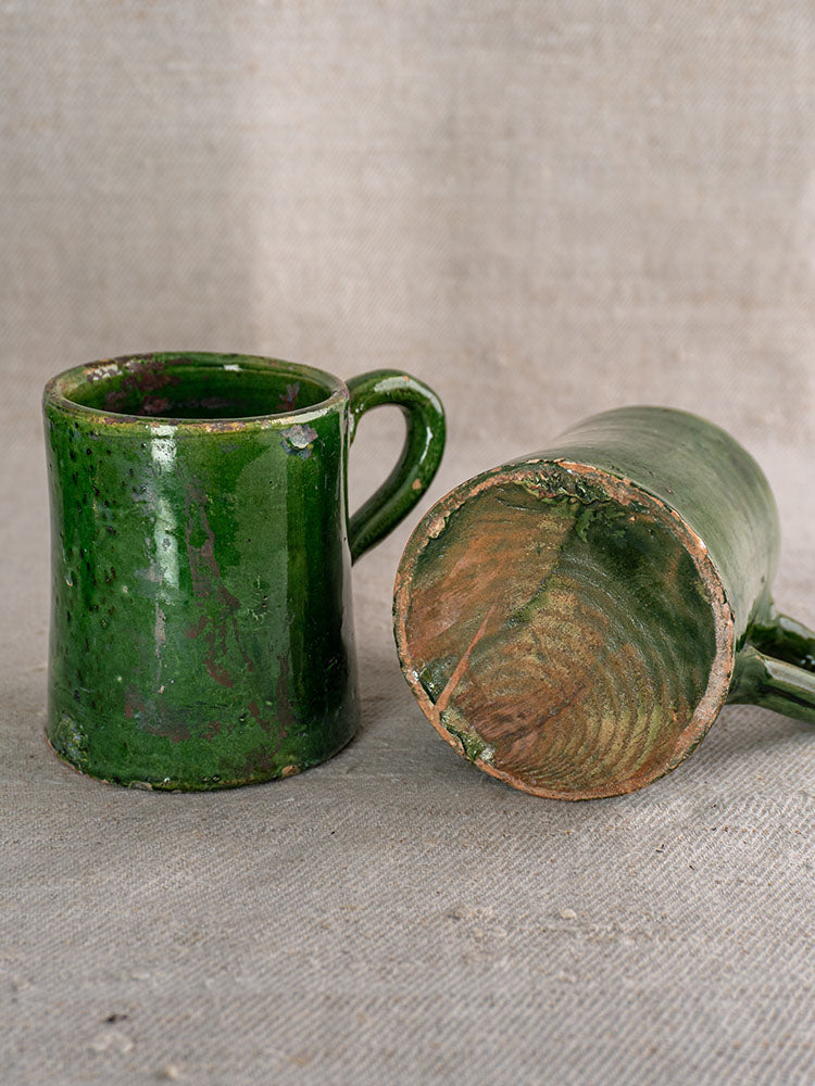 Two rustic French green glazed pottery mugs, one upright, one on its side revealing worn unglazed base, on beige fabric.