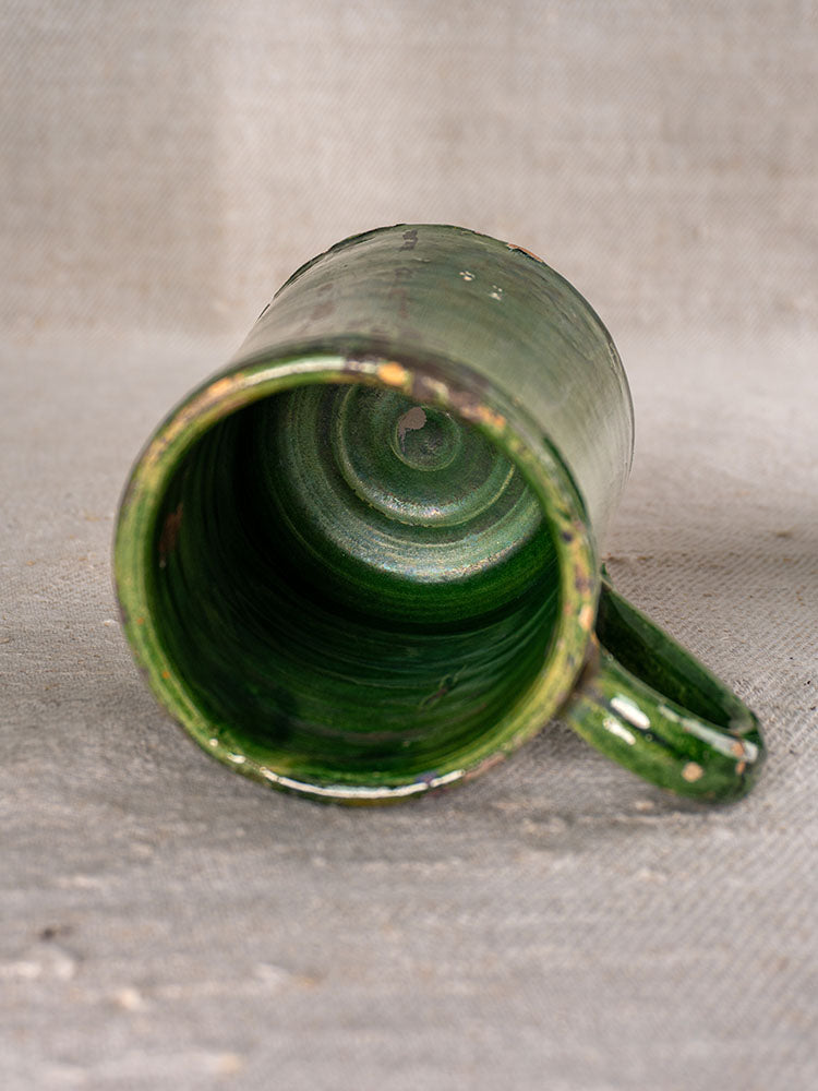 French green glazed pottery piece, handle visible, interior exposed on textured neutral fabric background.