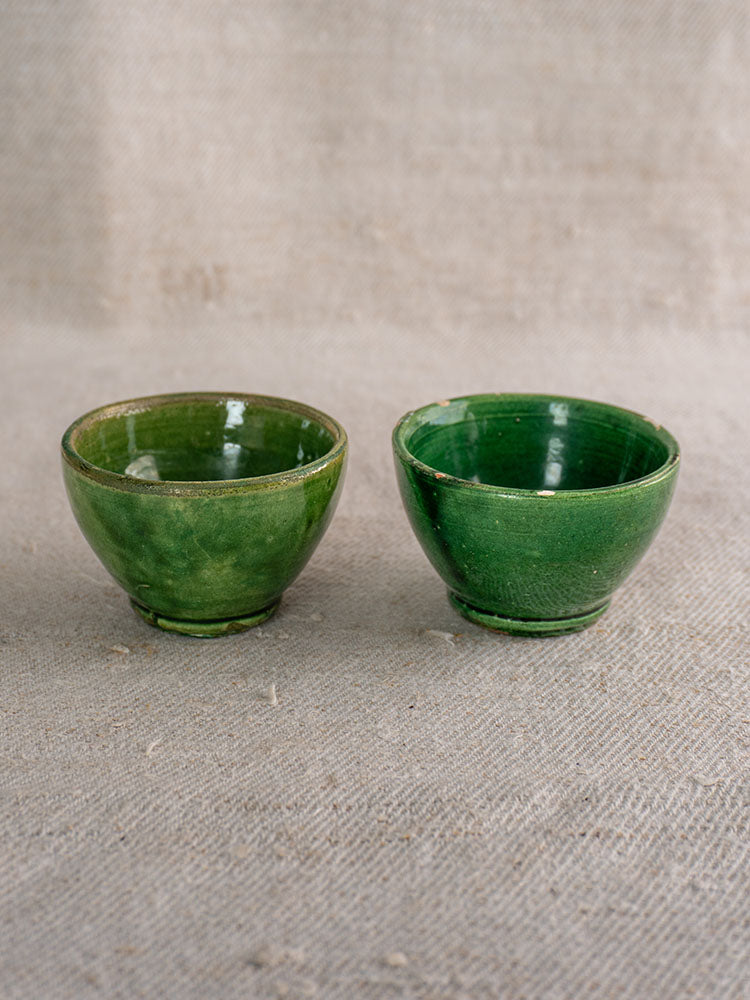 Two small French green glazed bowls with unique shapes and glossy finish displayed on beige textured fabric.