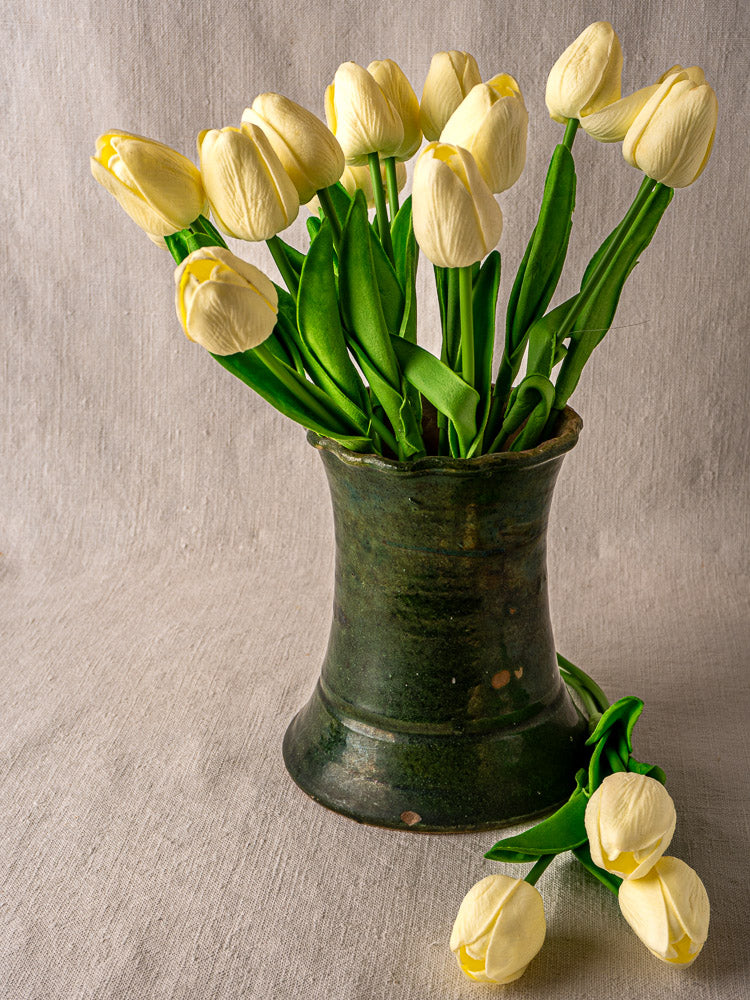 Antique French green glazed terracotta vase with pale yellow tulips on neutral fabric background.