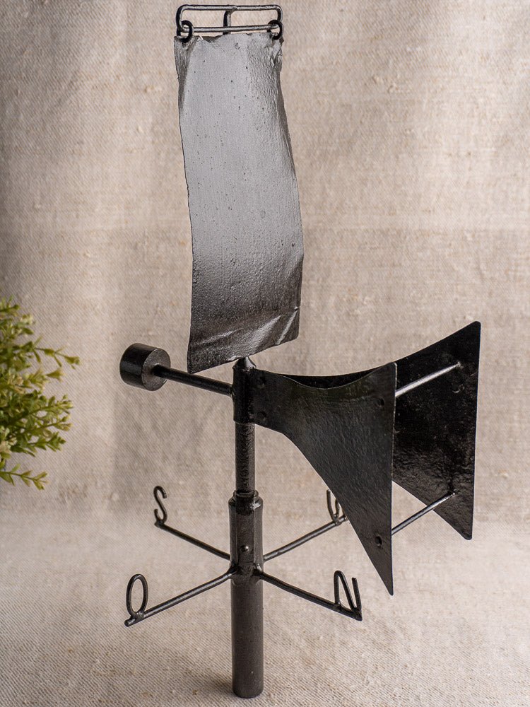 Antique French painted iron weathervane with black directional markers, displayed on neutral fabric with greenery.