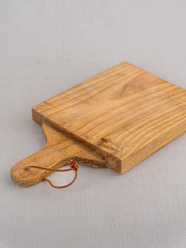 Mid 20th century French oak bread board with handle and leather loop, shown on light gray fabric.