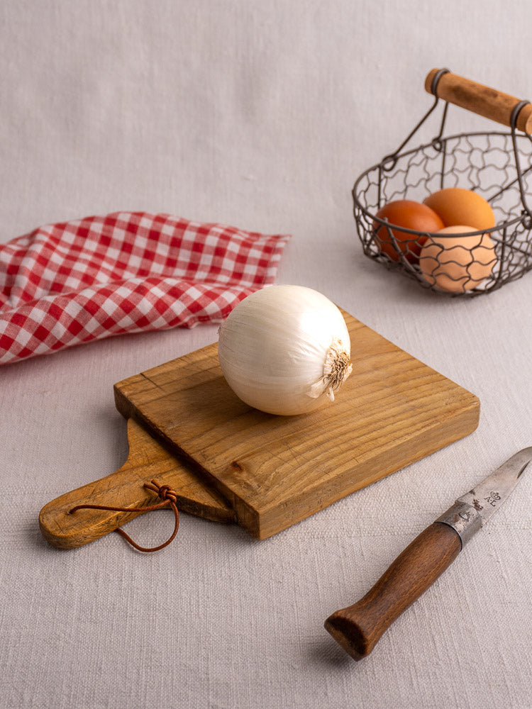 Mid 20th century vintage French bread board with whole white onion, red checkered dishtowel, eggs, and knife nearby.