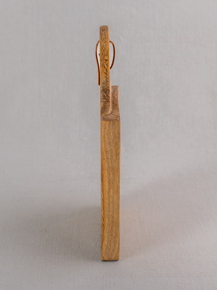 Vintage French bread board from the mid-20th century, upright on a light gray surface, leather hanging loop at top.