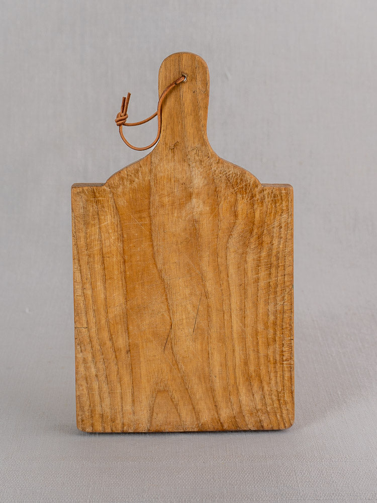 Mid 20th century French oak bread board with handle and leather loop shown upright on a neutral gray background.