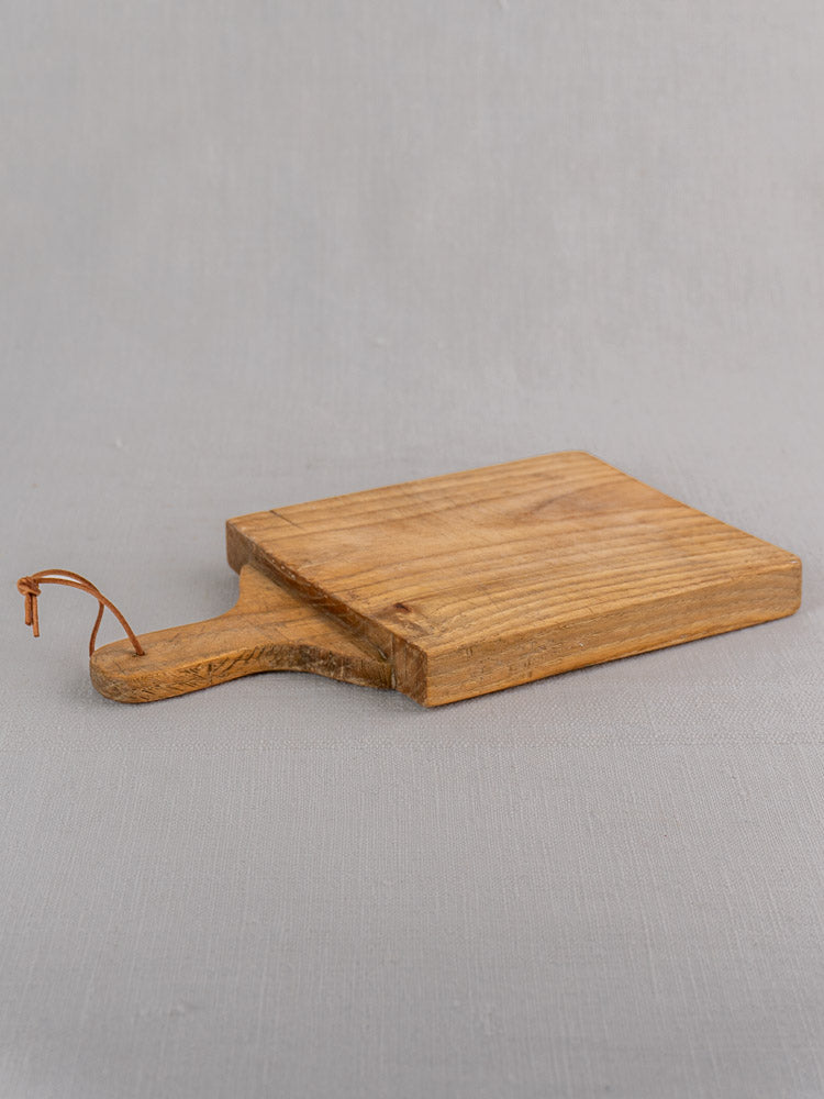 Small rectangular oak bread board with handle and leather loop, inspired by antique French style, on light gray surface.