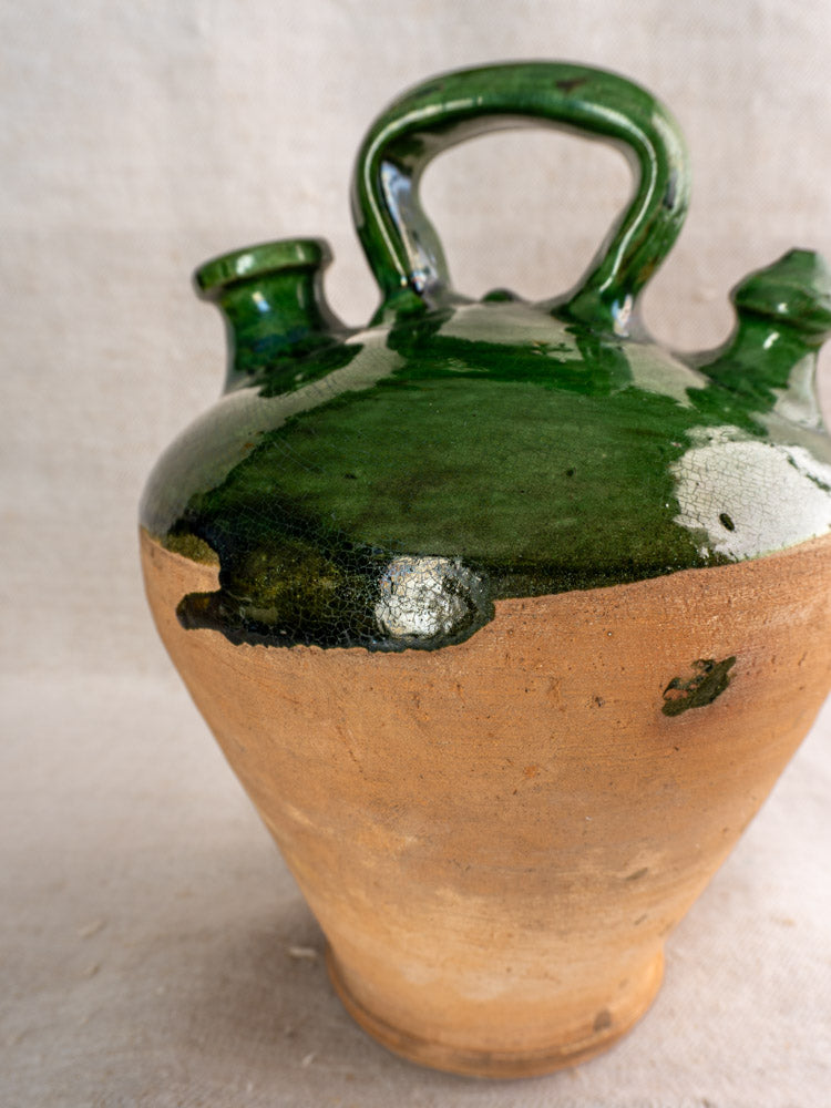 Antique French gargoulette jug with emerald green glazed top, unglazed brown base, two spouts, handle; plain background.