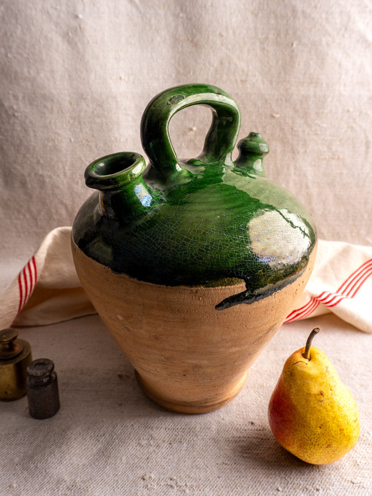 Antique French gargoulette with emerald green glaze, displayed on textured fabric beside a yellow pear and metal containers.