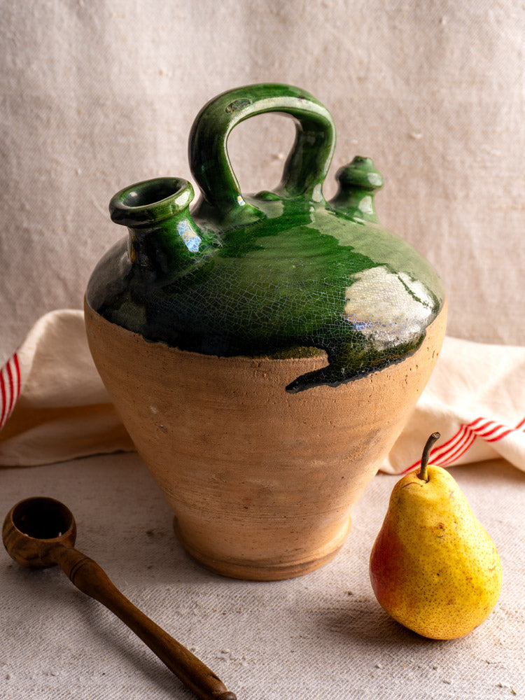 Antique French gargoulette with emerald green glaze, shown with a yellow pear and pipe on textured fabric.