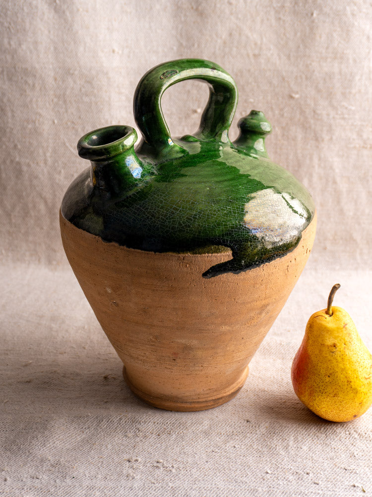 Antique French gargoulette with emerald green glaze, circa 19th century, beside a yellow pear on beige fabric.