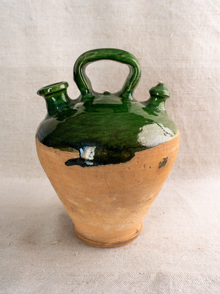Antique French gargoulette with emerald green glaze, glazed top, handle, and dual spouts on neutral textured fabric.