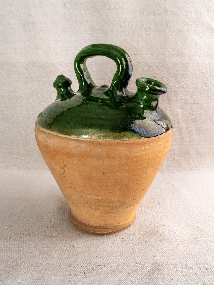Antique French gargoulette with tan base, emerald green glazed top, two spouts, and handle on neutral fabric background.